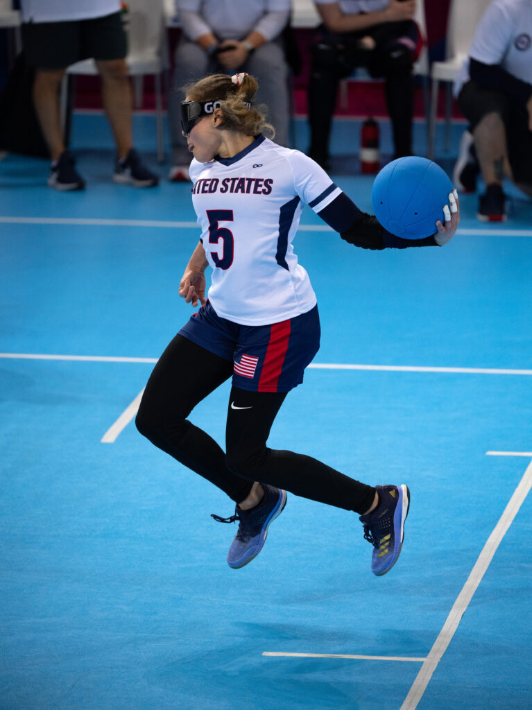 With both feet off the ground, Amanda Dennis spins in the air as she prepares to unleash a throw during the 2019 Parapan American Games in Lima, Peru. She is wearing a white jersey with number 5 and the words "United States on the front, as well as blue shorts with the American flag on the left thigh over black tights.