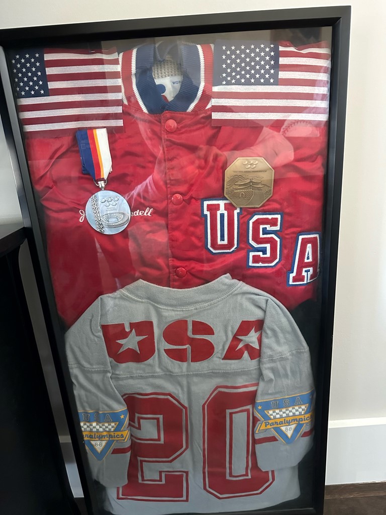 A frame showing a red USA jacket, silver medal, belt buckle and with #20 USA jersey belonging to Jerry Windell.