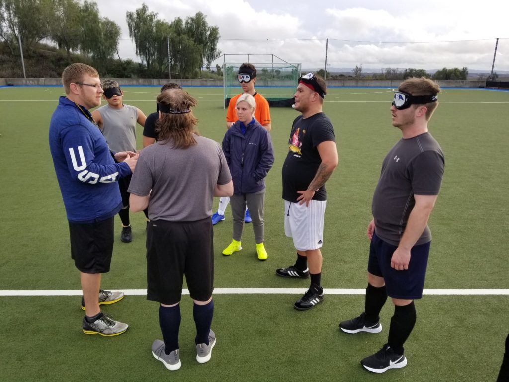 Athletes wearing eyeshades listen to coach Tim Taylor in the middle of the soccer field