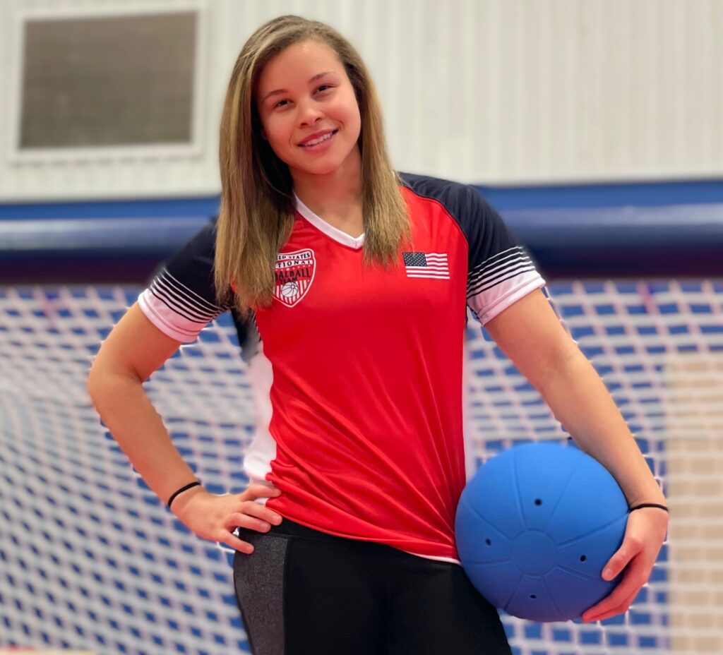Amanda Dennis poses for a photo in front of a goalball goal. She is wearing a red USA Goalball National Team shirt with an American flag on the chest and is cradling a goalball in her left hand.