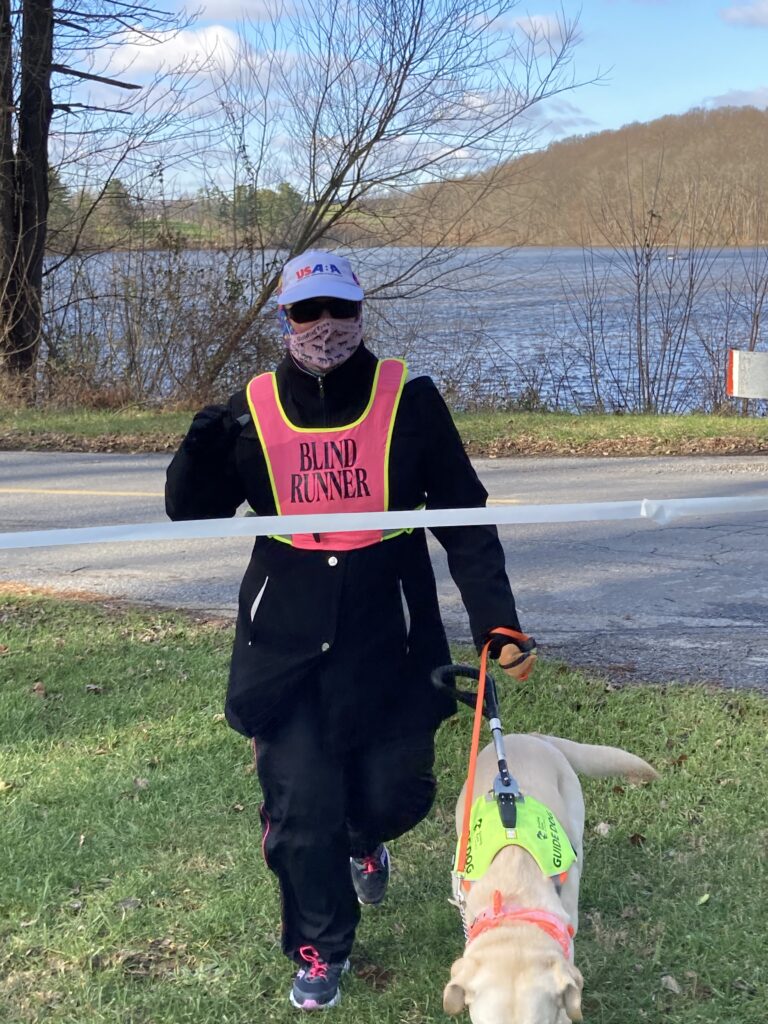 Wearing her USABA hat, a mask and Blind Runner vest, Cindy Lou Altman runs through the tape with her guide dog Jada to finish her 5k run on National Blind Running Unity Day. In the background are trees and a lake.