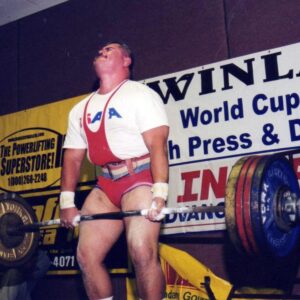 Cody Colchado is shown executing a dead lift with the barbell across the front of his thighs.