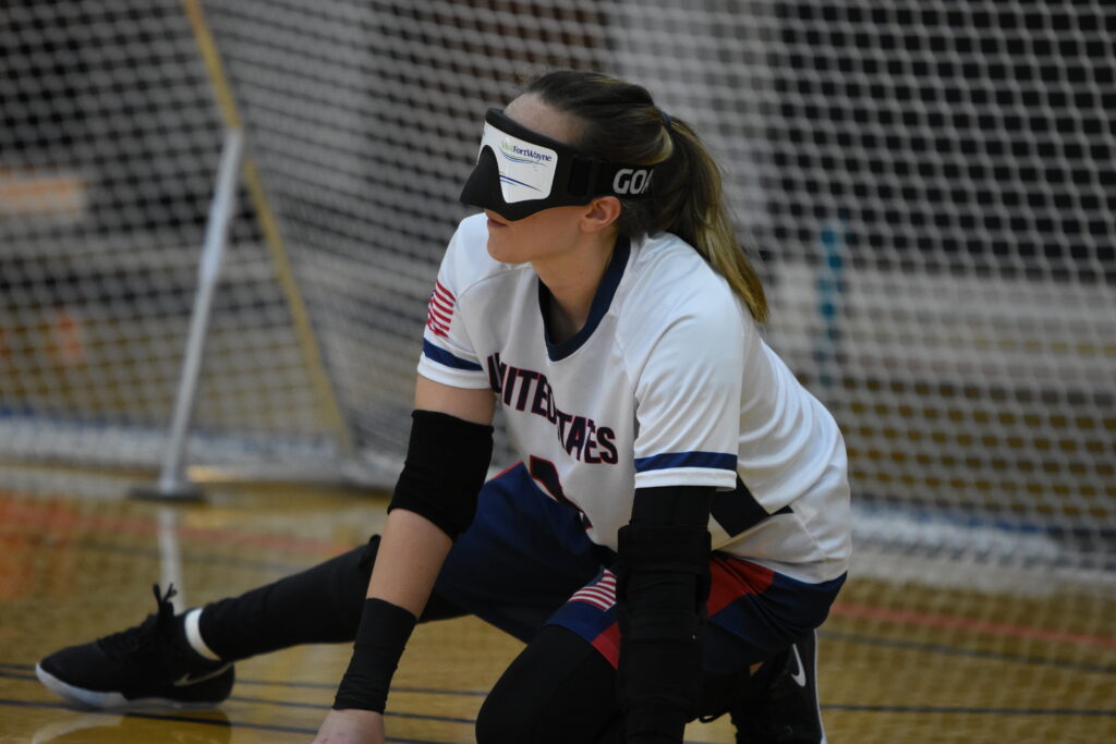 Wearing a white USA jersey and eyeshades, Eliana Mason crouches on the floor in front of her goal, ready to defend an opponent's throw.