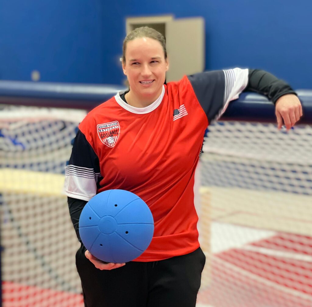Lisa Czechowski poses in front of a goalball goal with her left arm resting on the crossbar. She is wearing a red USA Goalball jersey and holding a blue goalball in her right hand.