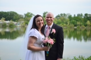 Daryl and his wife Melissa pose in front of a lake on their wedding day. Melissa is wearing a white wedding dress and veil, and Daryl is wearing a dark suit coat, white dress shirt and pink tie.