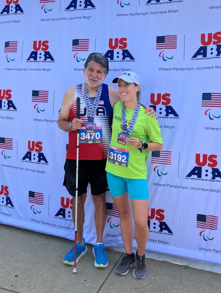 Ramiro Contreras and guide Caitlin Nelson pose for a photo with their medals in front of a USABA backdrop