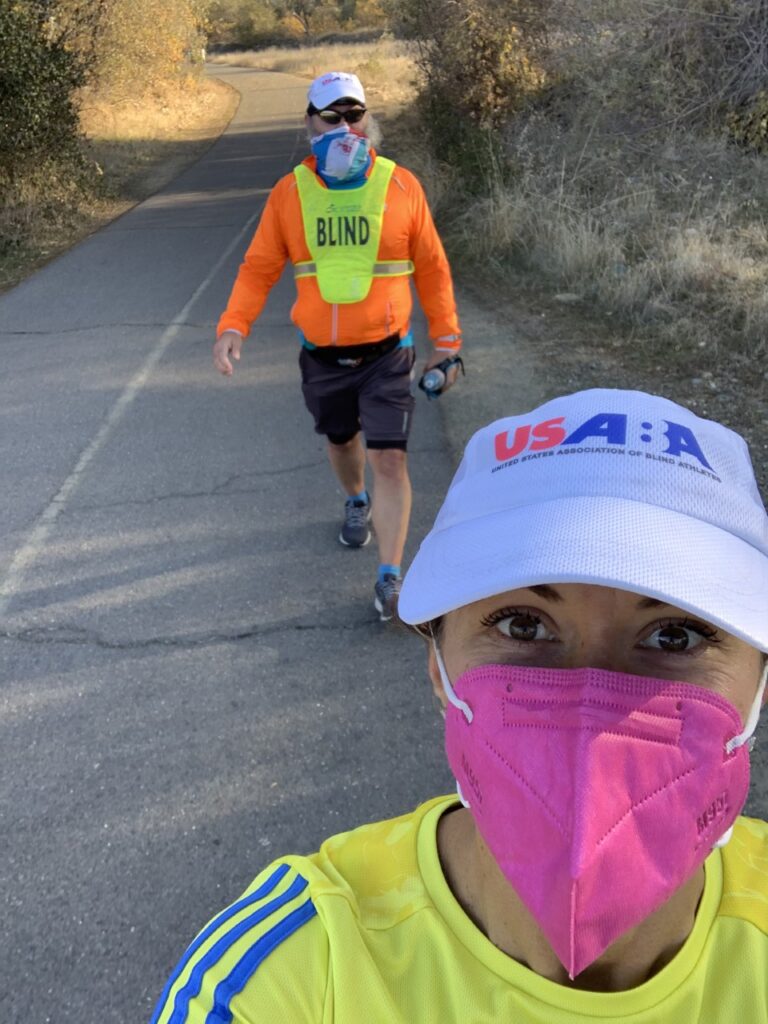 Rob Sanchas' guide takes a selfie of the two runners on a path. Rob is in the background wearing his yellow bib with the word "BLIND" on it. Both runners are wearing masks and USABA running hats.