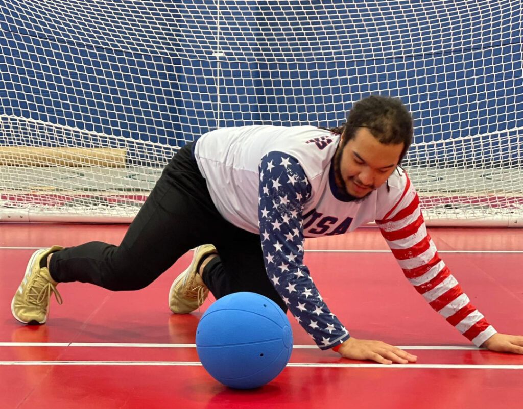 Sean Walker on his hands and knees in front of a goalball goal with a blue goalball in front of him. He is wearing a USA jersey and black pants.