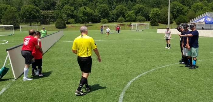 A referee wearing a yellow shirt and black shorts officiates a blind soccer scrimmage.
