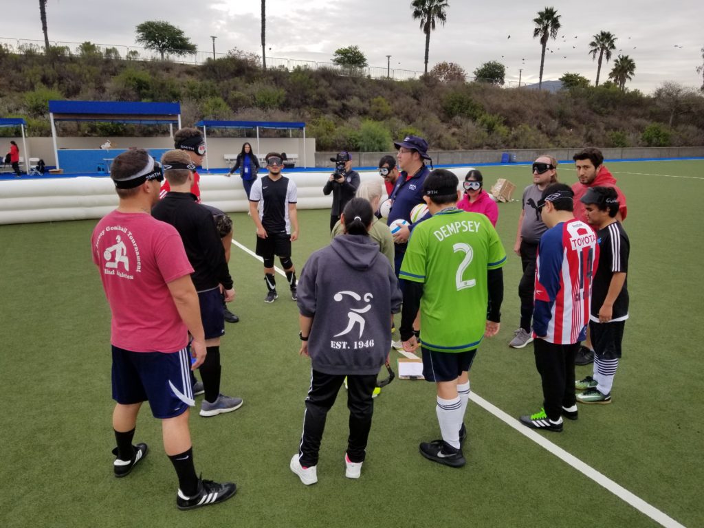 Athletes attending the talent identification camp gather around coach Tim Taylor at midfield of the soccer pitch in Chula Vista, Calif. The athletes are wearing eyeshades and an array of soccer jerseys and competition gear. Palm trees can be seen in the background.