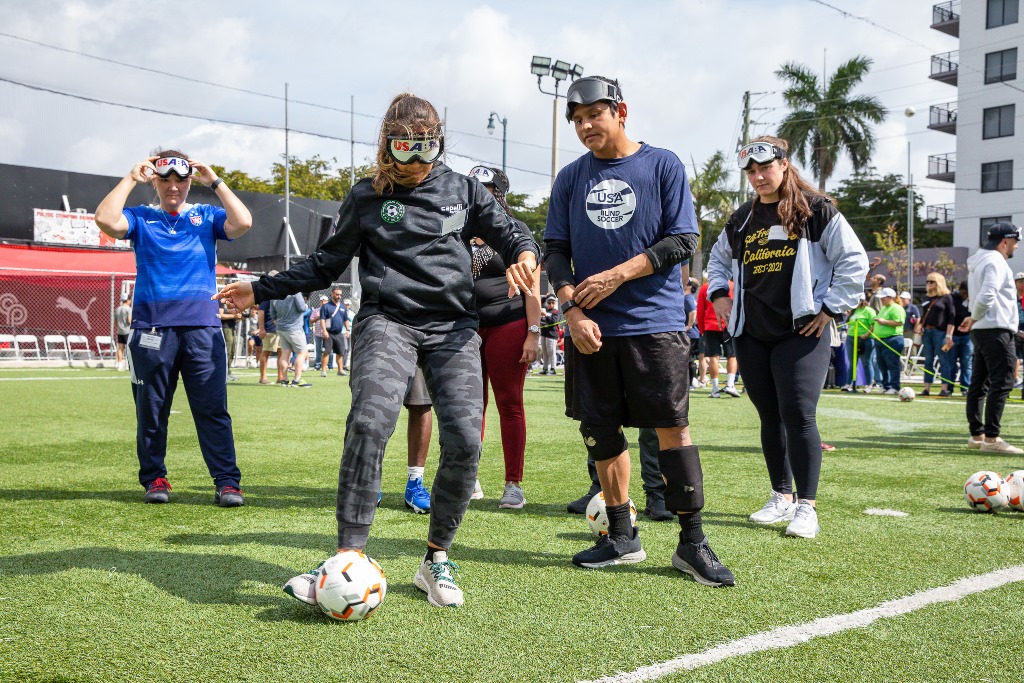USA Blind Soccer athlete Ricky Castaneda provides instruction to participant Erika Deetjen as she kicks a ball while wearing eyeshades.