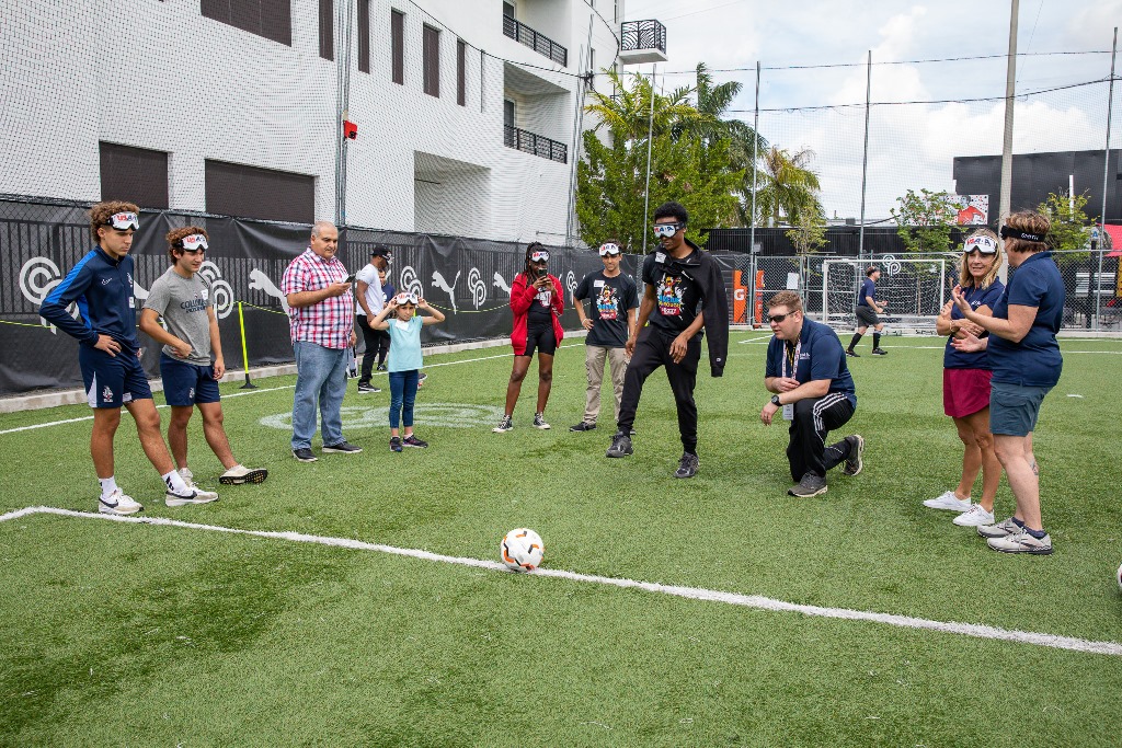A participant executes a penalty kick under the guidance of Coach Tim Taylor as other participants look on.
