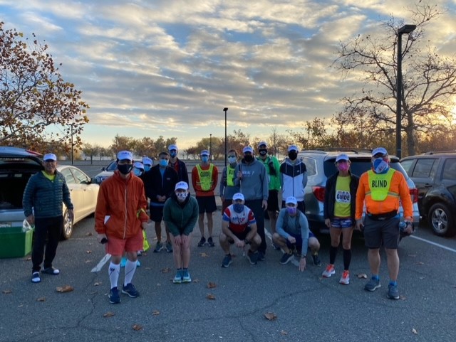 A group of 16 runners and guides wearing masks pose for a group photo in a parking lot. Some are wearing GUIDE or BLIND bibs.