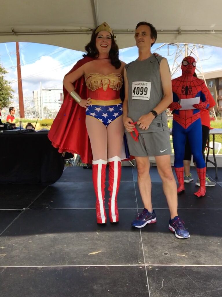 William poses with a woman dressed as Wonder Woman while a man dressed as Spiderman stands in the background during the CASA Superhero 5k. Proceeds of this race go to help protect children who have been abused.