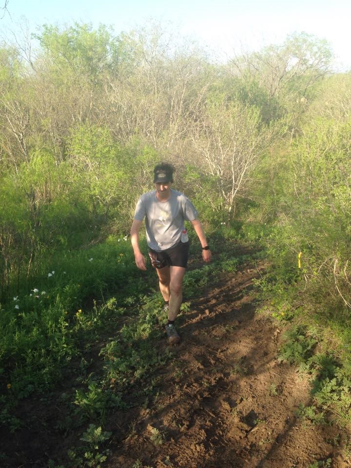 William Greer is shown running on a muddy trail through green trees and bushes during the Prickly Pear 50k. For this race, he came in third place in his age group.
